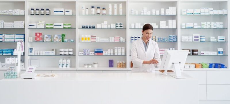 Moderna apoteka sa ฤistim belim policama, na kojima su poreฤani razliฤiti medicinski proizvodi i lekovi. Farmaceut u belom mantilu stoji iza pulta i koristi kompjuter, pruลพajuฤi profesionalnu pomoฤ kupcima.