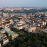 Panoramski pogled iz vazduha na centar Beograda, prikazujući istorijske zgrade, moderne građevine i ušće Save i Dunava u pozadini, pod vedrim nebom.