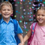 Dvoje srećne dece, dečak i devojčica, drže se za ruke i smeše dok idu u školu, okruženi šarenim konfetama koje padaju oko njih. Oboje nose školske rančeve