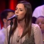 Danica Crnogorčević nastupa na sceni, pevajući u mikrofon, u tradicionalnoj srpskoj nošnji, sa horom u pozadini