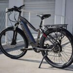 Električni bicikl siva boje sa integrisanom baterijom, opremljen sa zadnjim nosačem za teret i prednjim amortizerom, parkiran napolju.