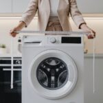 Osoba meri širinu veš mašine pomoću metra u modernoj kuhinji.