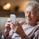 Starija žena sedi na sofi i smeši se dok gleda u mobilni telefon, u pozadini topla i prijatna atmosfera dnevne sobe.