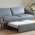 Elegantni sivi trosed na razvlačenje sa modernim dizajnom, idealan za dnevni boravak, postavljen na drvenom podu sa dekorativnim biljkama u pozadini.