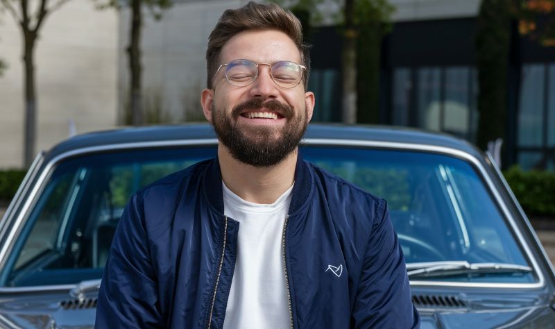 Nasmejan muลกkarac sa bradom i naoฤarima stoji ispred klasiฤnog automobila, uลพivajuฤi u trenutku