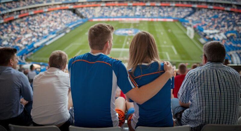 Zaljubljeni par u dresovima sedi na stadionu, grleฤi se dok gledaju fudbalsku utakmicu.