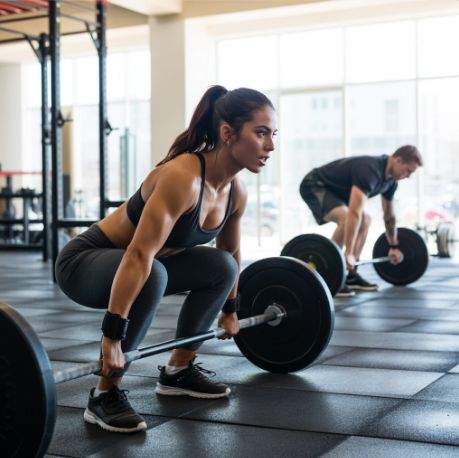 ลฝena u teretani izvodi mrtvo dizanje sa ลกipkom, dok muลกkarac u pozadini takoฤe trenira sa tegovima.