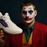 Nikola Jokić kao Džoker, drži svoju košarkašku patiku sa vibrantnim dizajnom, u crvenom odelu i sa šminkom Džokera na licu