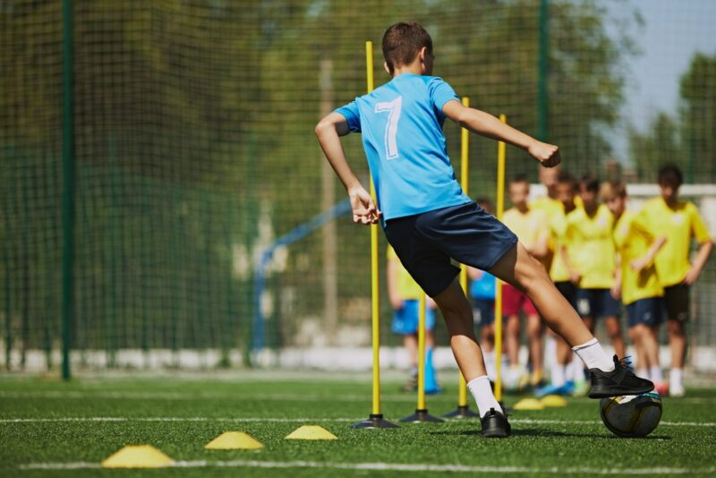 Dečak u plavoj sportskoj opremi trenira vođenje lopte između čunjeva na fudbalskom terenu