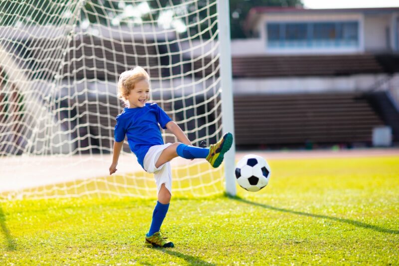 Mali dečak u plavoj fudbalskoj opremi šutira loptu ispred gola na fudbalskom terenu