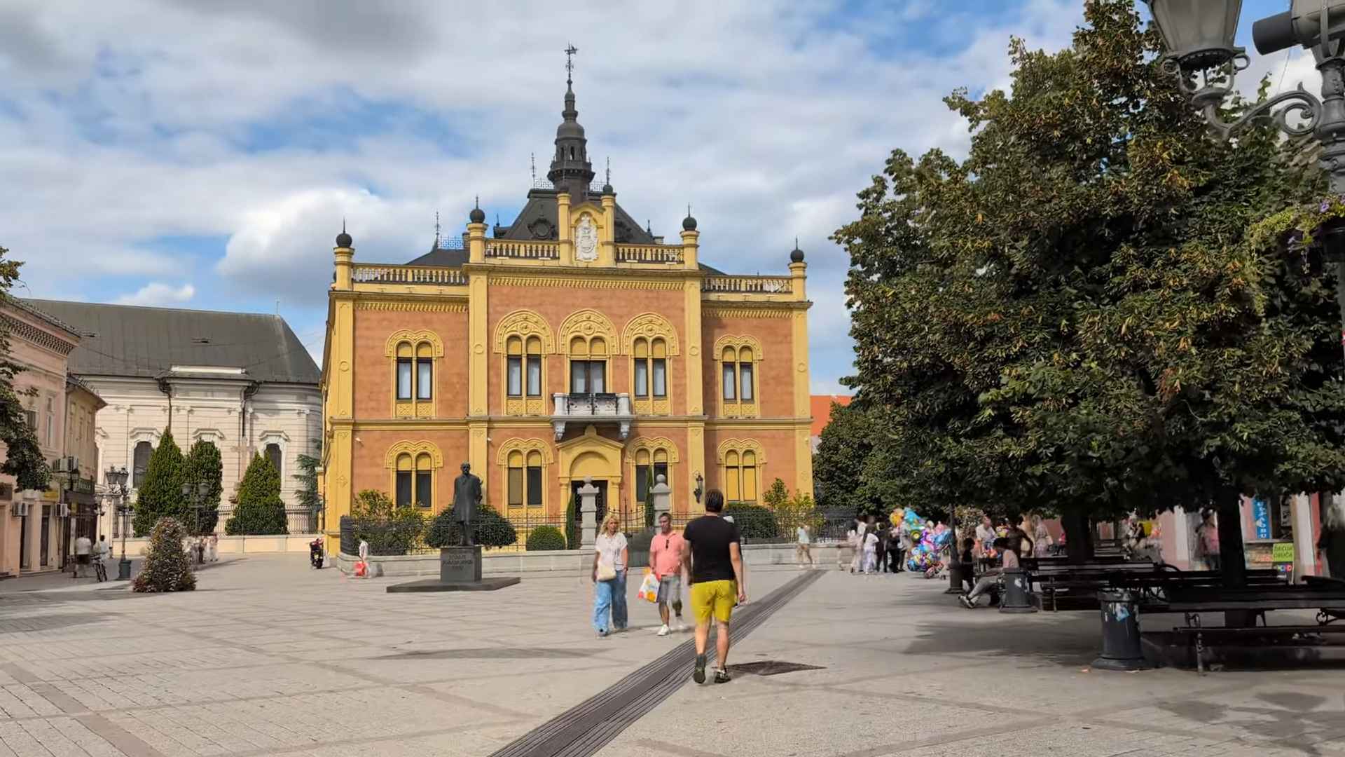 Trg u centru Novog Sada sa istorijskom zgradom i prolaznicima.