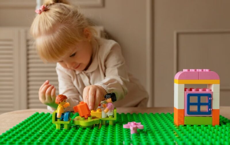 Plavokosa devojčica pažljivo slaže šarene LEGO kocke i figurice na zelenoj podlozi, pored male kućice napravljene od kockica