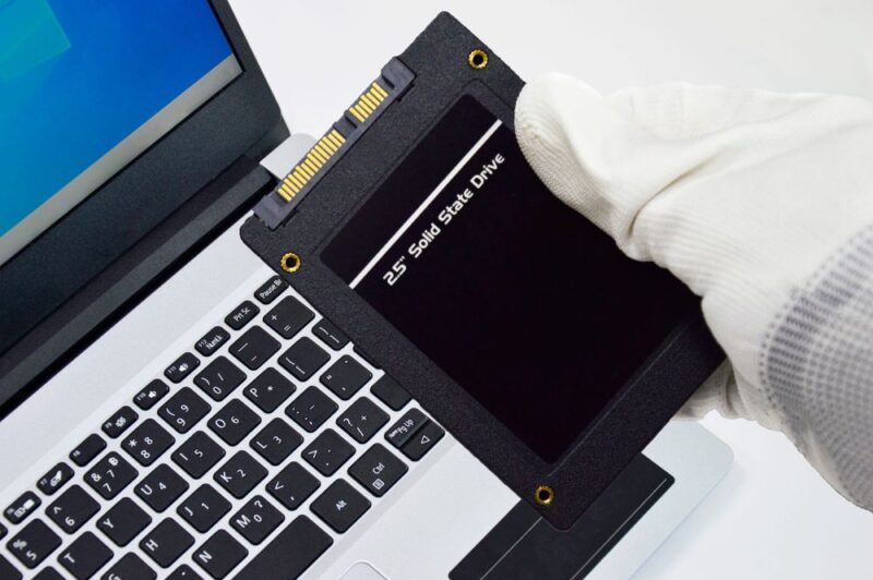 Ruka u beloj rukavici drži 2,5-inčni SATA SSD disk iznad tastature laptopa, kao primer nadogradnje računara